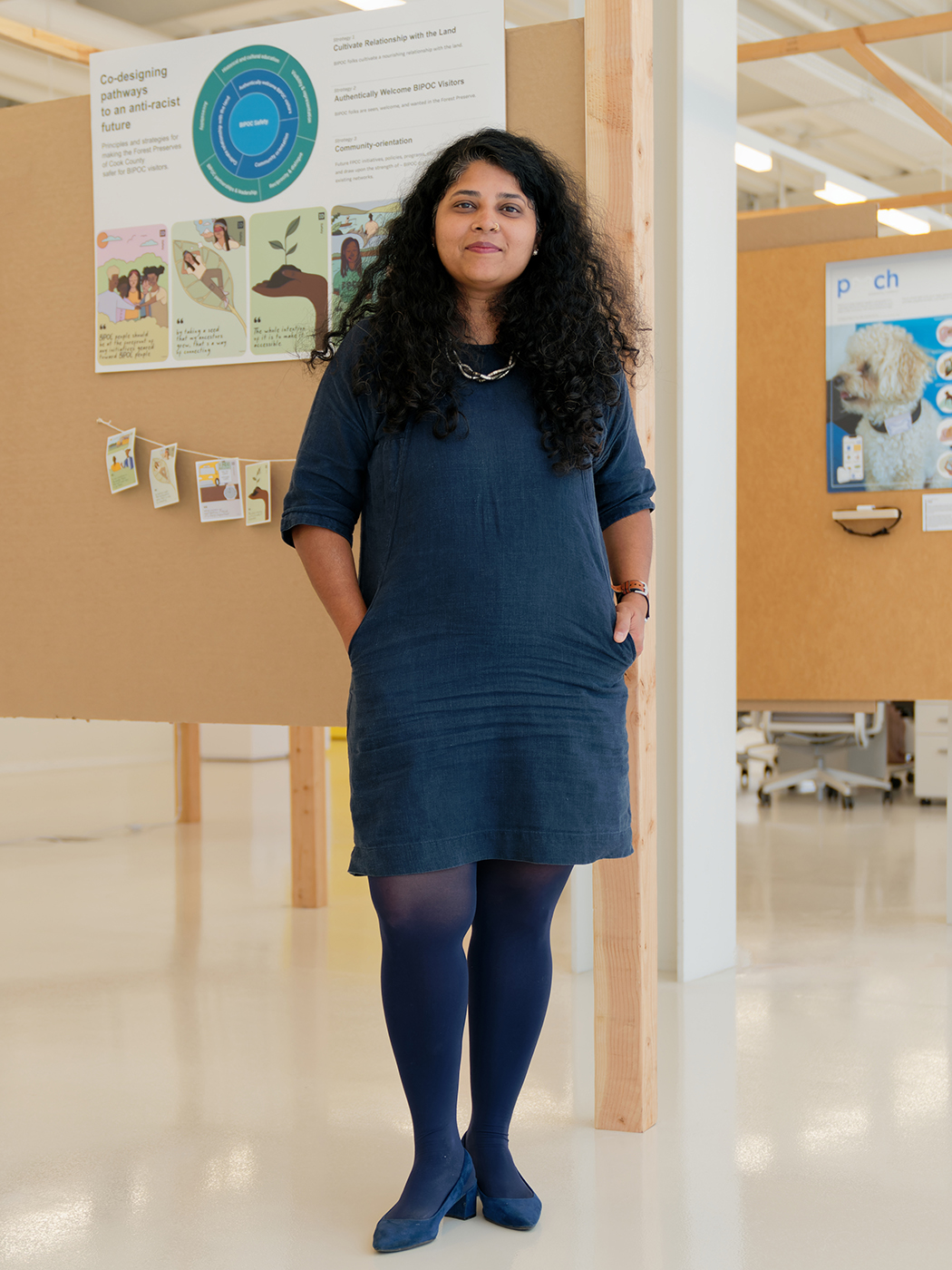 Shilpi Kumar, a Brown woman with long dark curly hair and a blue dress, stands in an ID studio